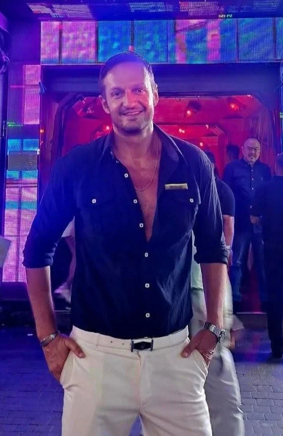 Yannis Pagiannidis at a club entrance in Pattaya at night, in a navy shirt and white trousers, hands in his pockets